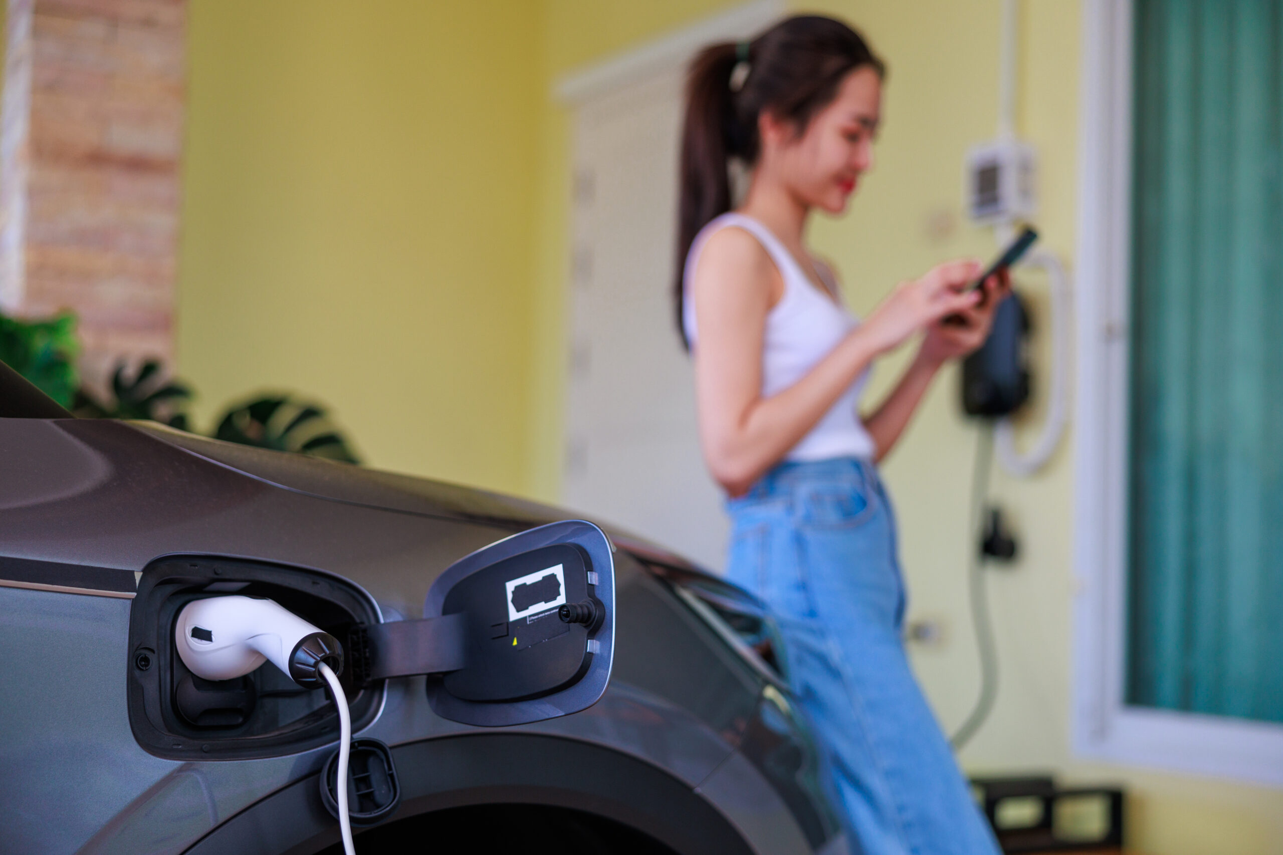 Sustainable Living: Asian Woman Plugs in Electric Charger for Car at Home, Embracing Eco-Friendly Transportation