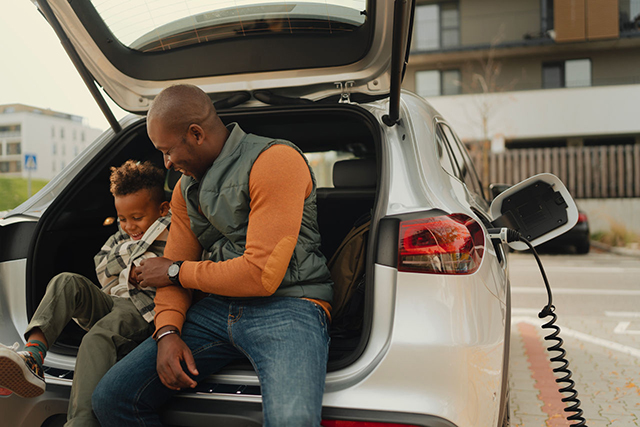 Multiracial father with his son sitting in car trunk and waiting while their electric car charging.