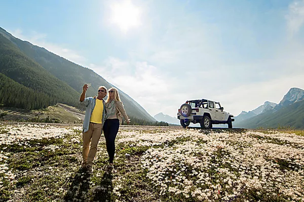 A couple takes a selfie in a flower-filled valley, with a Jeep parked behind them and mountains all around.
